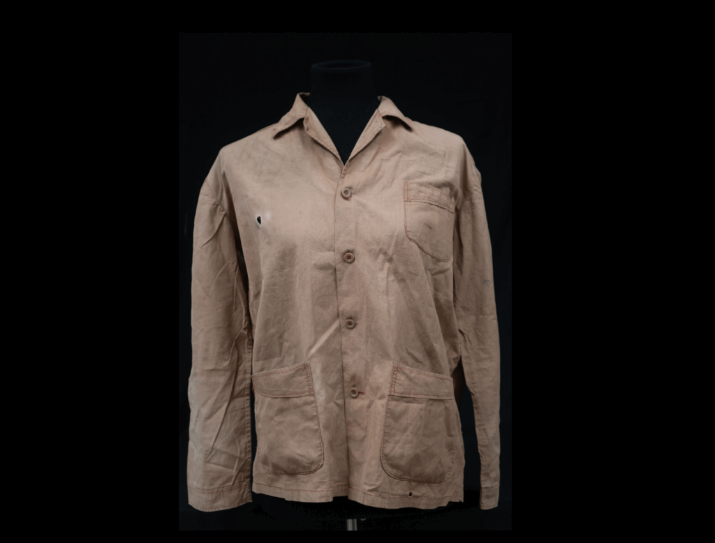 A beige, long-sleeved, cotton button-up men’s shirt with a visible bullet hole on the right side of the chest. The fabric is slightly wrinkled with signs of wear.